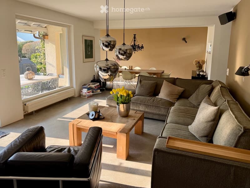 Modern living room with large windows offering a view, featuring a grey sectional sofa, wooden coffee table, leather armchair, dining area with chairs, and decorative items including a flower vase and books.