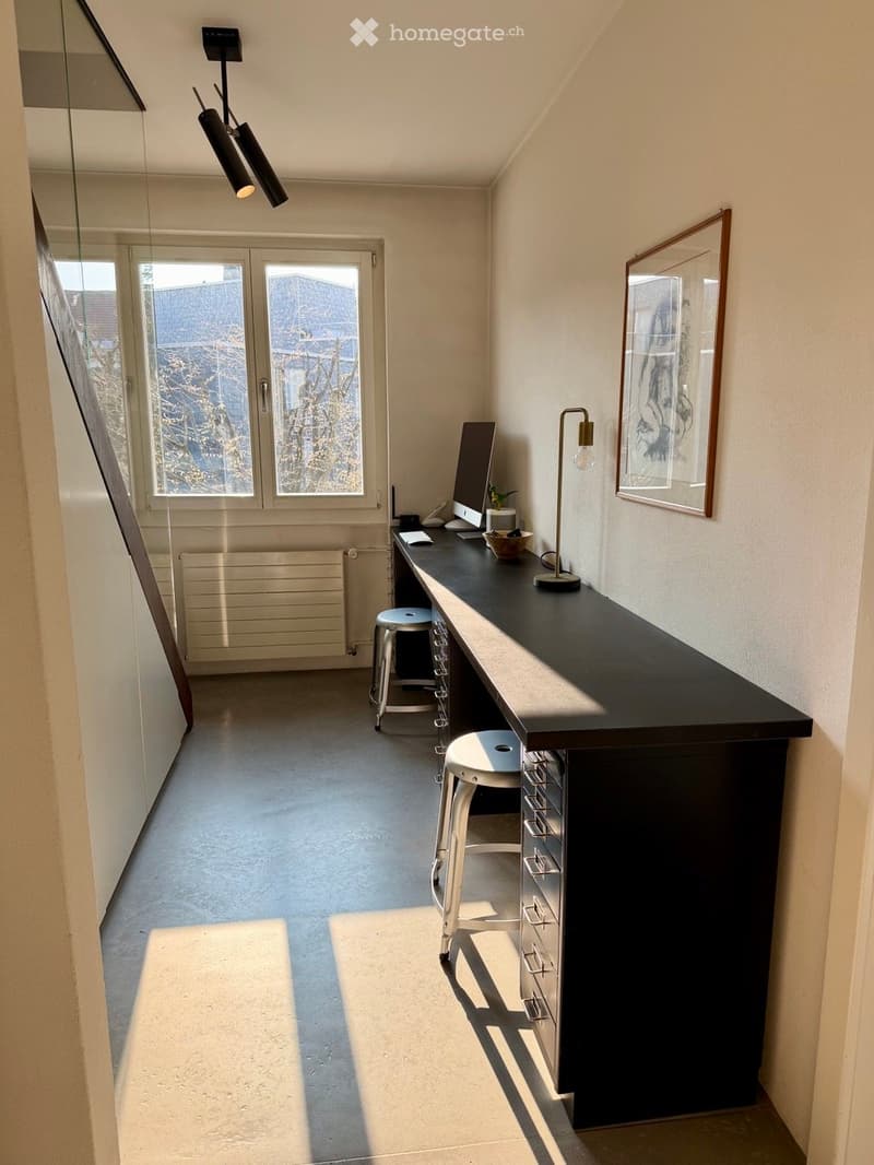 A modern, minimalist room with a black desk, two stools, a lamp, a framed picture, a computer monitor, a telephone, and a small plant. The room features large windows and a sleek, contemporary design.
