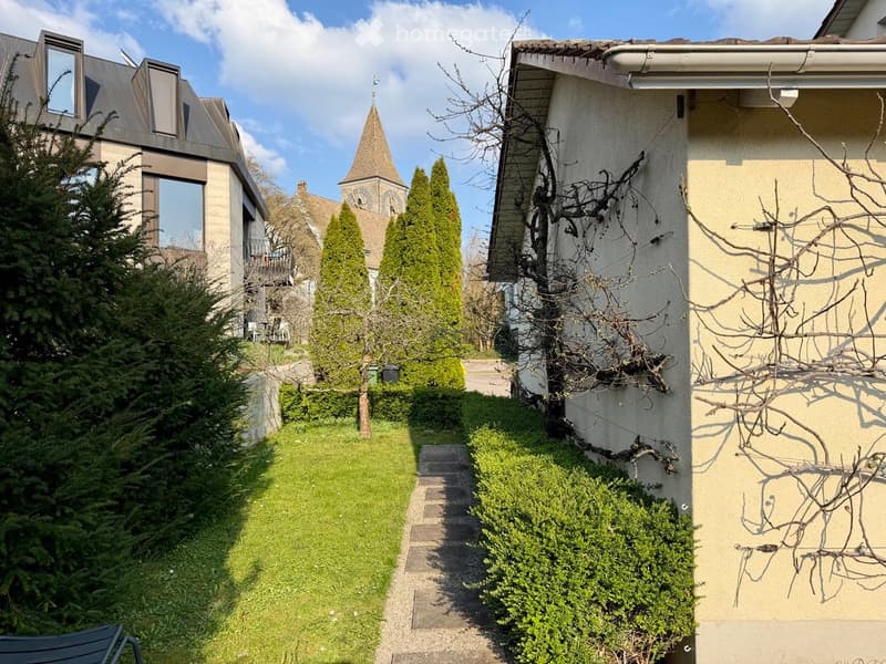 A well-maintained garden path leads to a house with a conical-roofed church visible in the background. The garden features trimmed hedges, a small lawn, and a few trees, including a conifer and a bare tree with branches leaning against the wall.