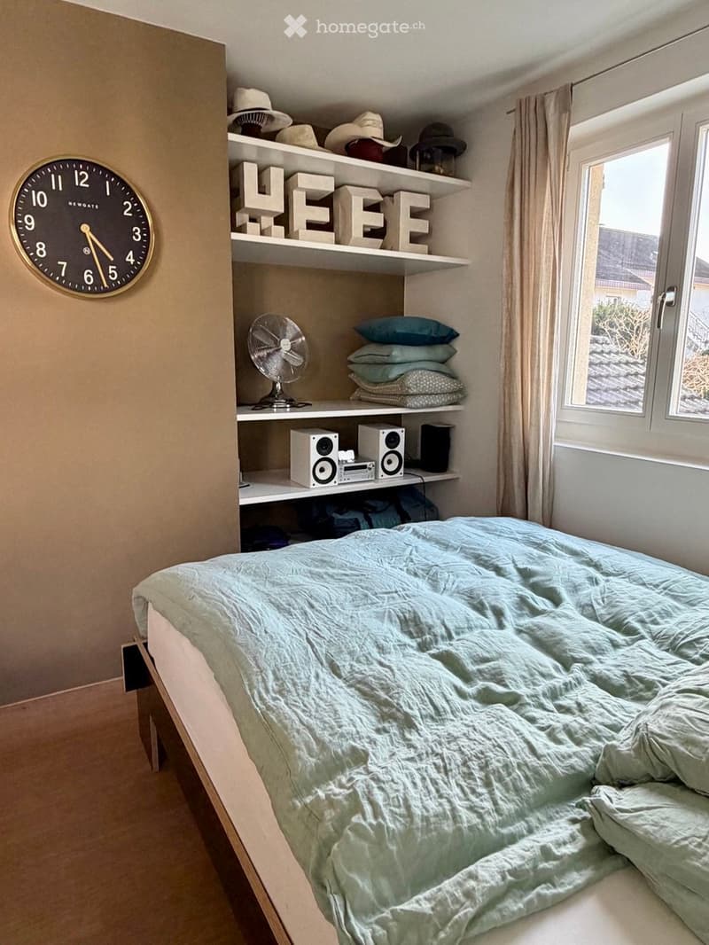 A cozy bedroom with a neatly made bed, a wall-mounted shelf holding hats, a fan, pillows, and audio equipment, and a large window with curtains.