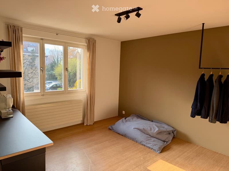 A furnished room with a wooden floor, a table with a sewing machine, a pillow on the floor, curtains, a coat rack with coats, and a TV mounted on the wall.