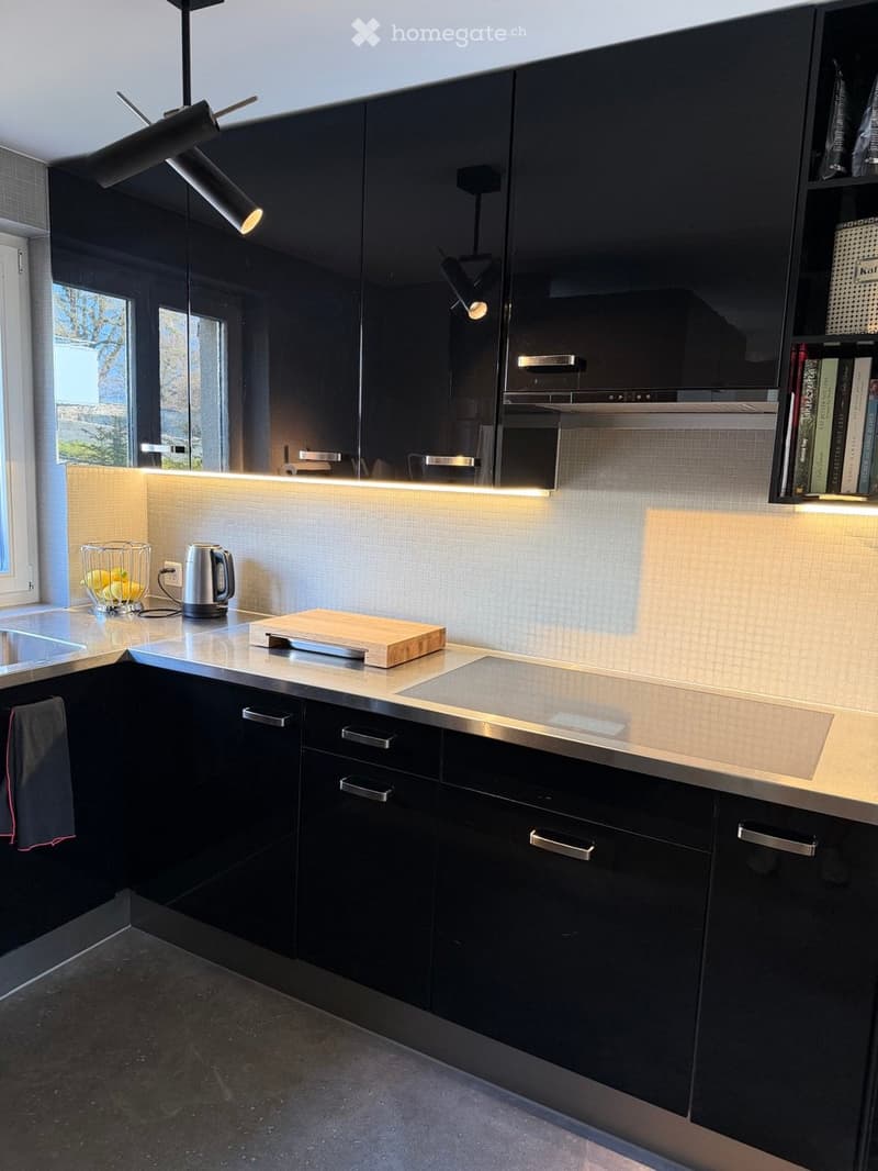 Modern kitchen with black cabinets, stainless steel appliances, and a sleek countertop. Features include a built-in oven, a fruit basket, and a wooden cutting board on the counter.