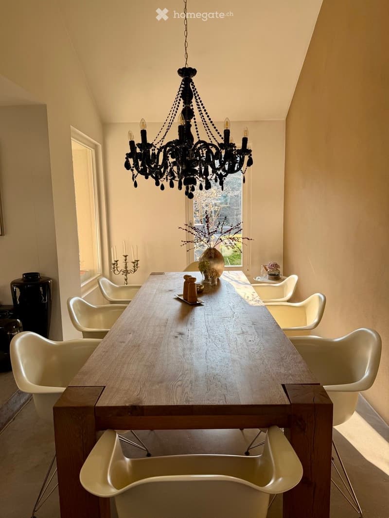 A modern dining room with a long wooden dining table surrounded by white chairs, a chandelier hanging from the ceiling, and a vase with dried flowers on the table.