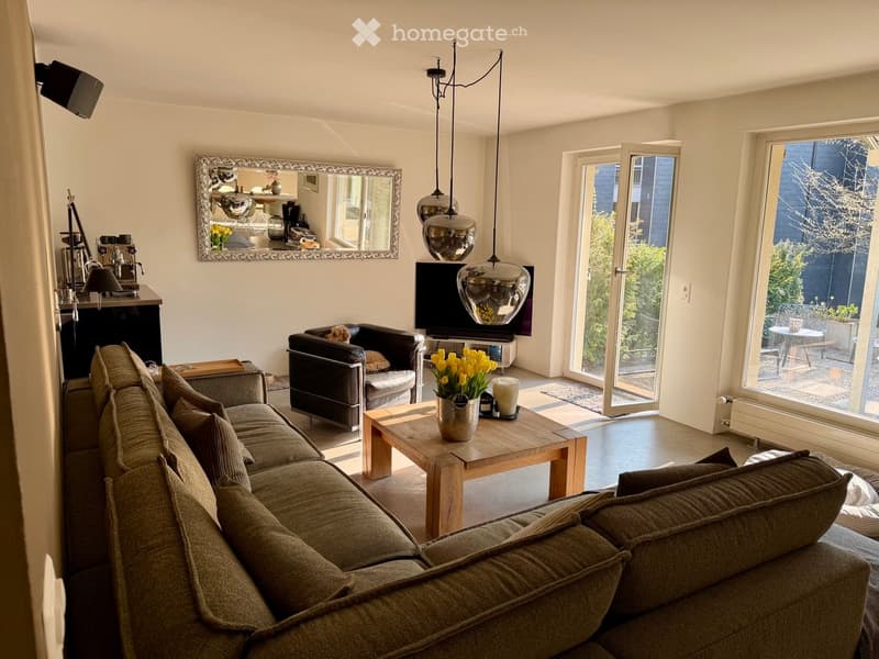 Bright living room with a grey sectional sofa, wooden coffee table, armchair, TV, and large mirror. Features pendant lights and a view of the garden through large windows and sliding doors.
