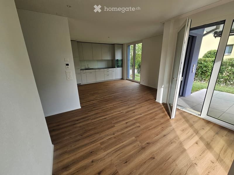 Open-plan space with wooden flooring, white walls, and large sliding glass doors leading to a patio. The kitchen area features white cabinets and a countertop.