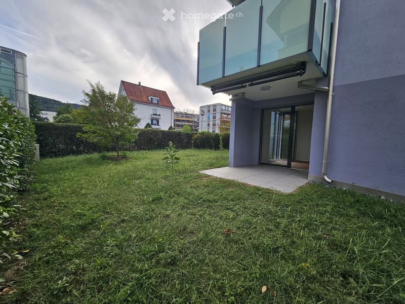 Lawn with a small tree, hedge, and a modern house with a balcony and glass doors.