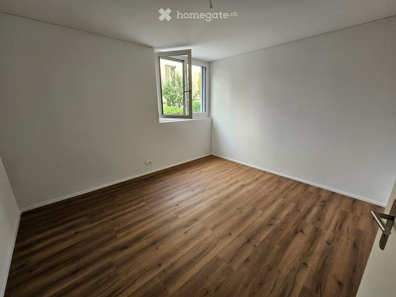 Empty room with wooden flooring, white walls, and a window with a view of greenery outside.