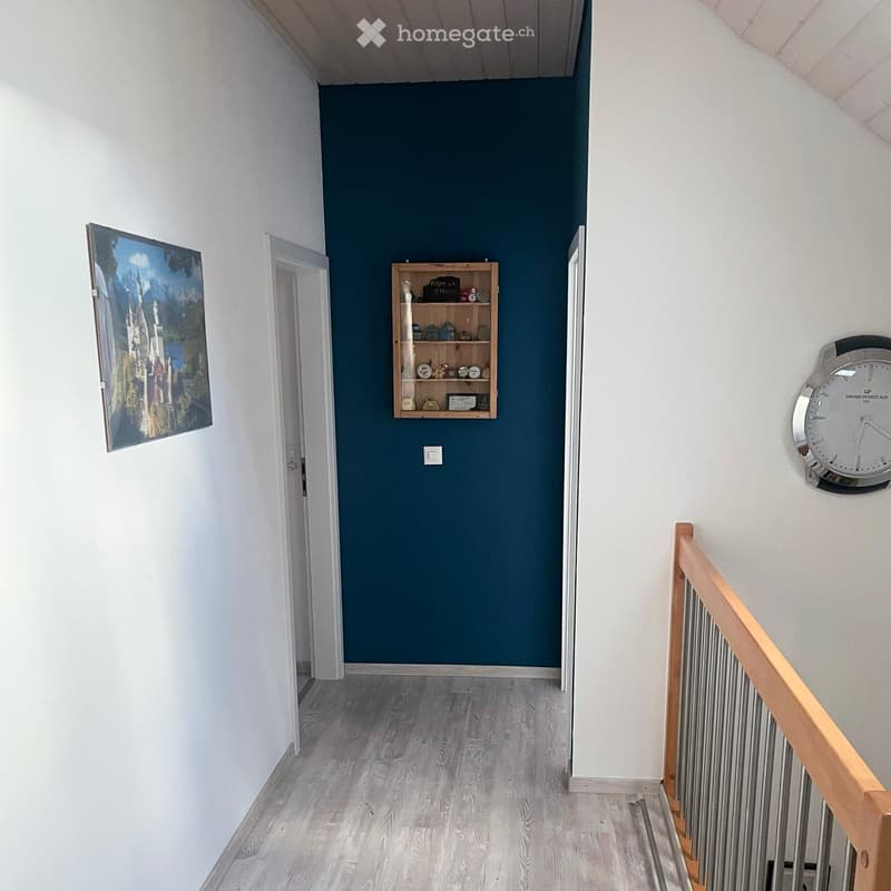 A narrow hallway with light wooden flooring, white walls on one side, and a dark blue accent wall on the other. Features include a small wooden shelf with decorative items, a large wall clock, and a painting on the left wall. A staircase with a wooden rai
