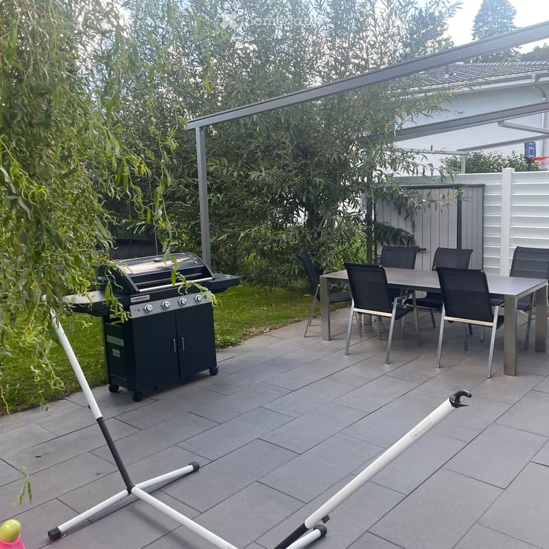 Outdoor patio with a grill, dining table, and chairs, surrounded by greenery and a covered area.