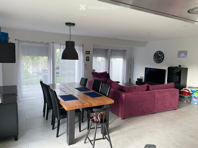 Open-plan living and dining area with a large wooden dining table, black chairs, a red sofa, a television, and a clock on the wall.