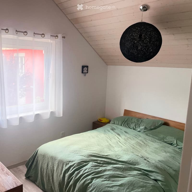 Cozy bedroom with a green duvet-covered bed, wooden nightstand, hanging spherical lamp, and sheer white curtains on the window.