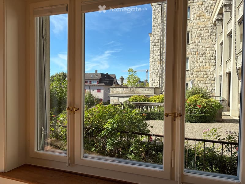 Open balcony with large windows overlooking a garden and cityscape, featuring greenery and stone buildings.
