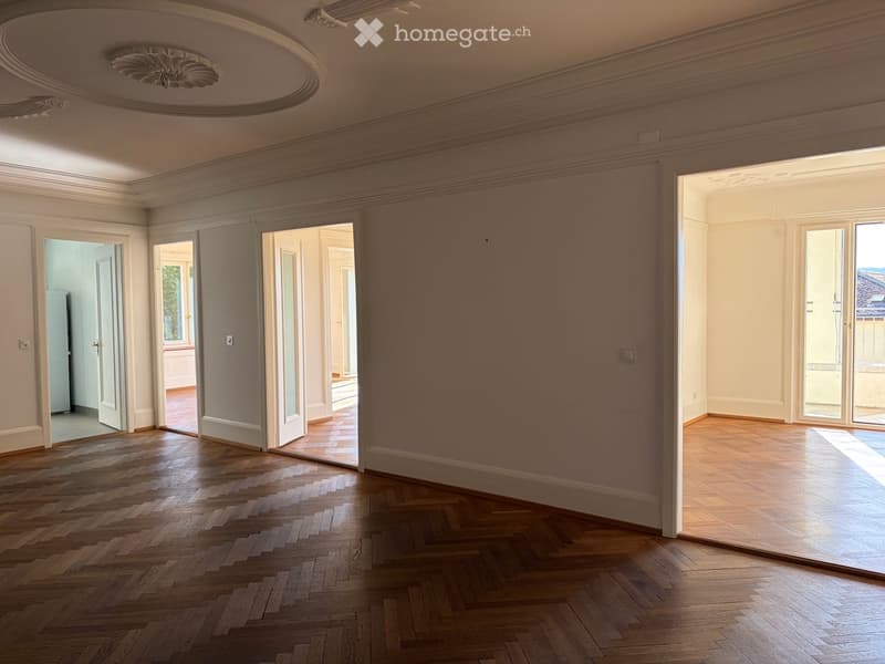 Empty room with wooden herringbone flooring, white walls, large windows, and open doors leading to other rooms.