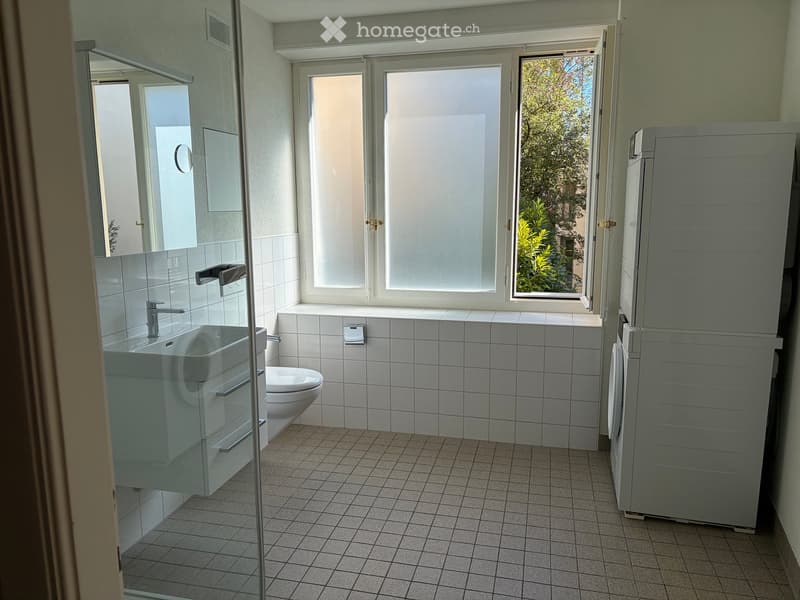 Modern bathroom with white tiled walls and floor, a sink with a chrome faucet, a toilet, a mirrored cabinet, and a large window letting in natural light.