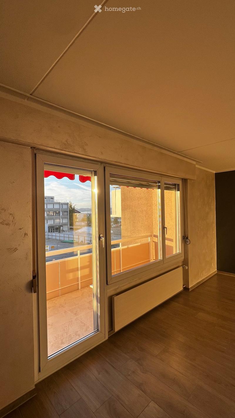 Empty room with wooden flooring, large window with open sliding doors leading to a balcony, radiator visible under the window.
