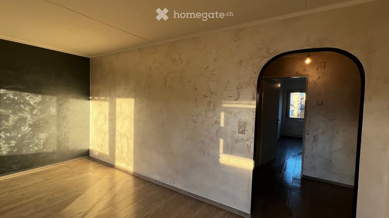 Empty room with wooden flooring, textured walls, and a large arched doorway leading to another space.