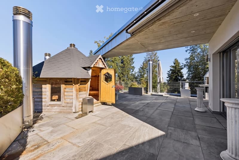 A modern outdoor patio area with a wooden sauna, stone tiles, and a covered section with wooden beams. The area includes a tall chimney, a closed umbrella, and surrounding greenery.