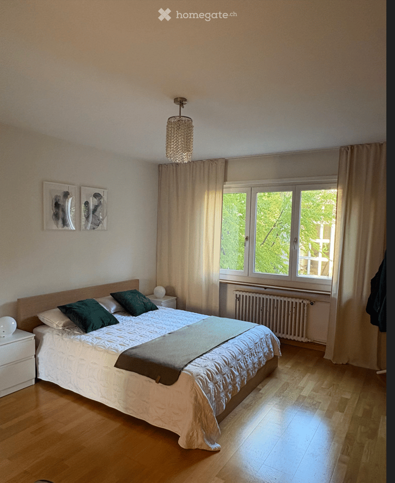 Bright bedroom with wooden flooring, large window with curtains, bed with green pillows and white comforter, nightstand with lamp, and wall-mounted picture frames.