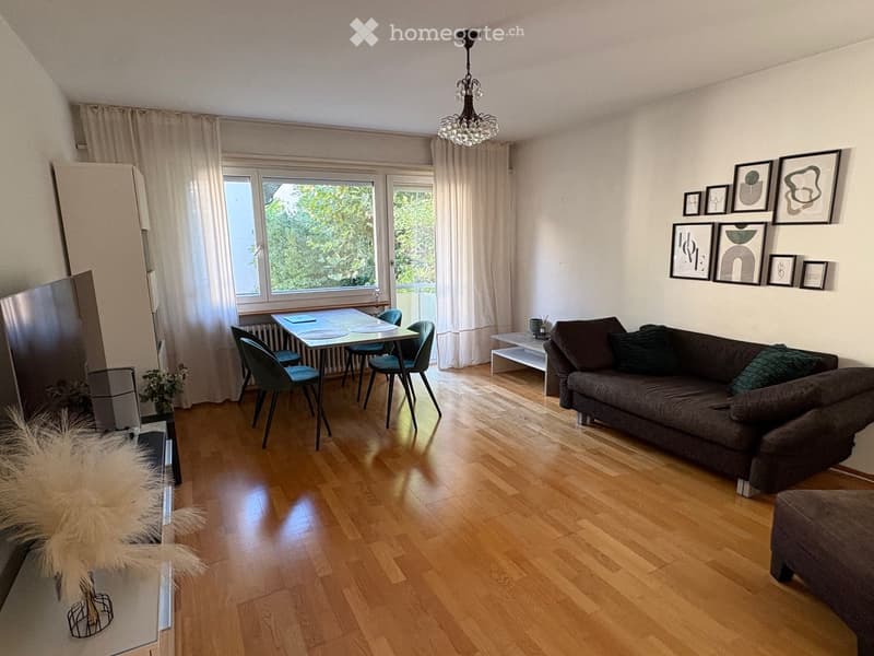 A modern living room with wooden flooring, a dark grey sofa, a small dining table with four chairs, a chandelier, and several picture frames on the wall.