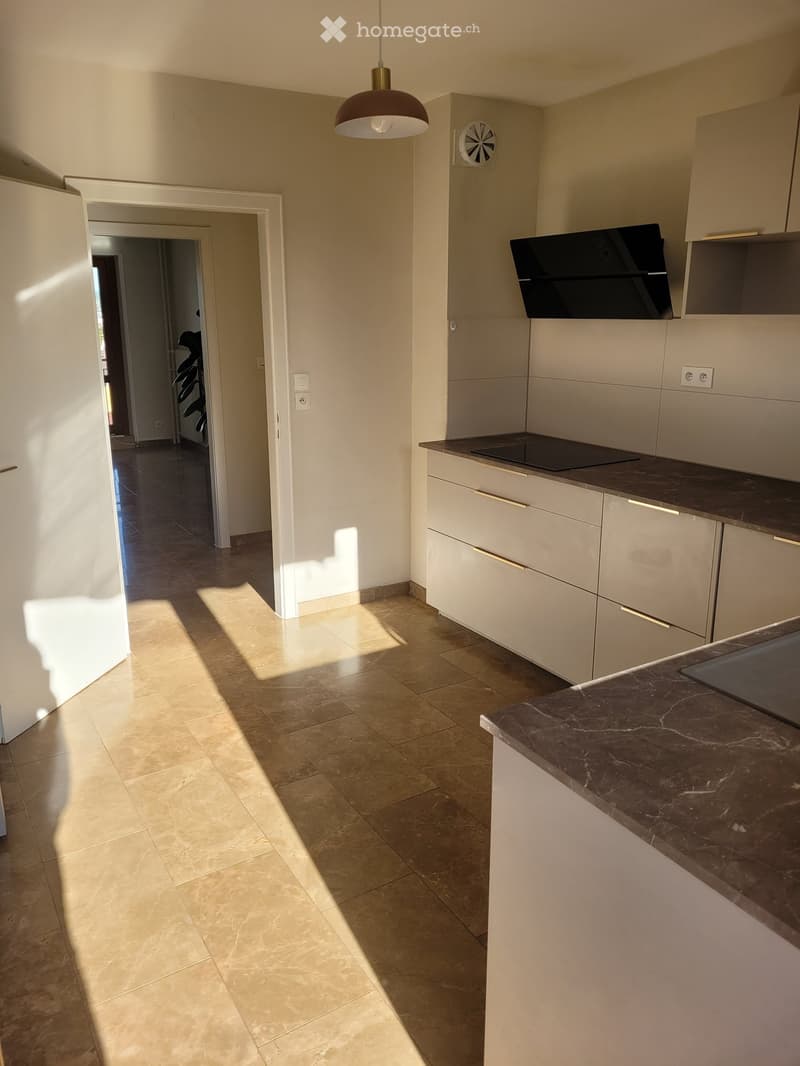 Modern kitchen with white cabinets, dark countertop, induction cooktop, and pendant light. Open space with tiled floor and natural light from adjacent room.