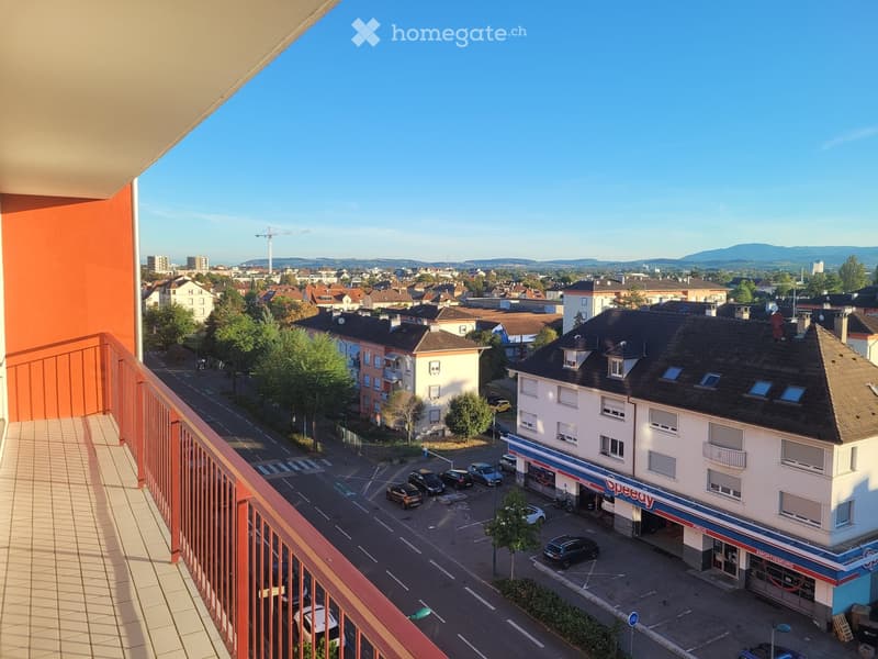 Balcony overlooking a street with buildings, parked cars, and a distant view of a cityscape and mountains.