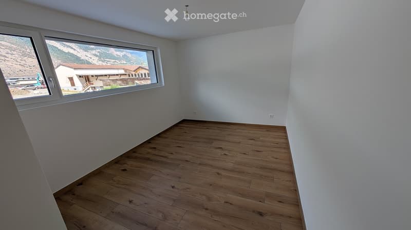 Empty room with white walls, wooden floor, large window, outside view of mountain and building.