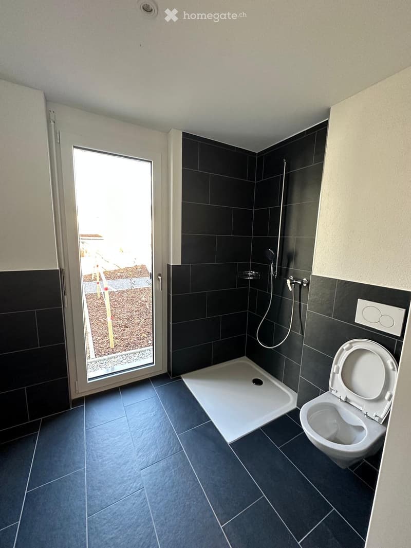 Modern bathroom with dark tiled walls and floor, a white shower stall, and a toilet with the lid up. A large window provides natural light and a view of the outdoor area.