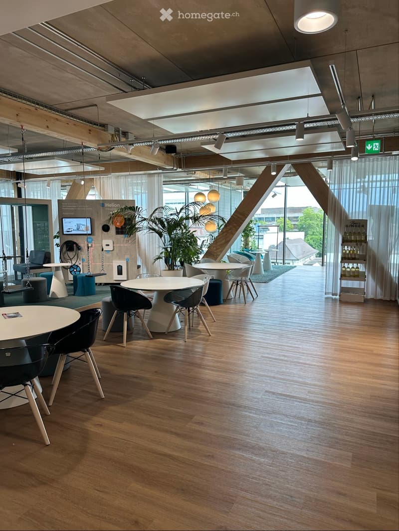 A conference room with wooden floors, white walls, round tables, chairs, green plants, a sign, monitors, and lamps. The room has large windows, wooden ceilings, and a glass window. Outside the window, there are trees and buildings.