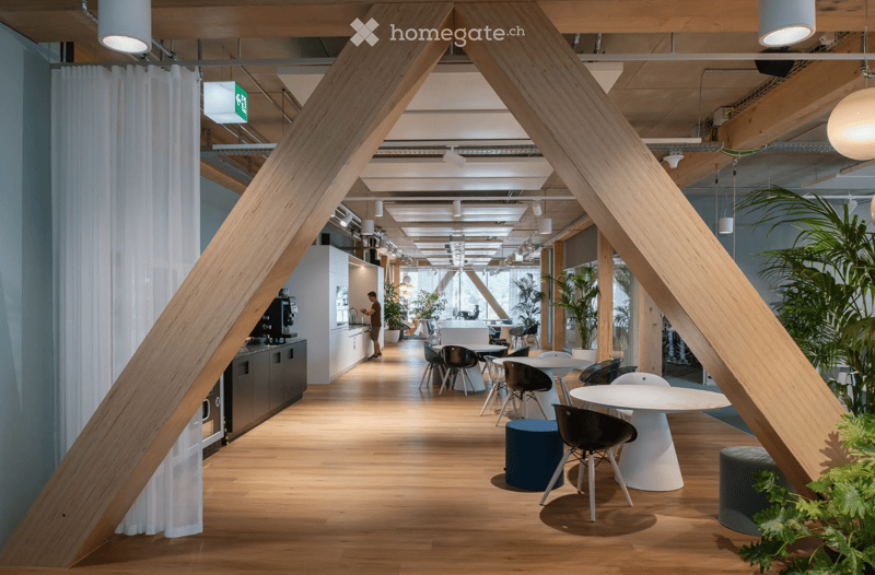 A large room with tables, chairs, wooden beams, wooden floor, plants, hanging lamps, white walls, white doors, curtains