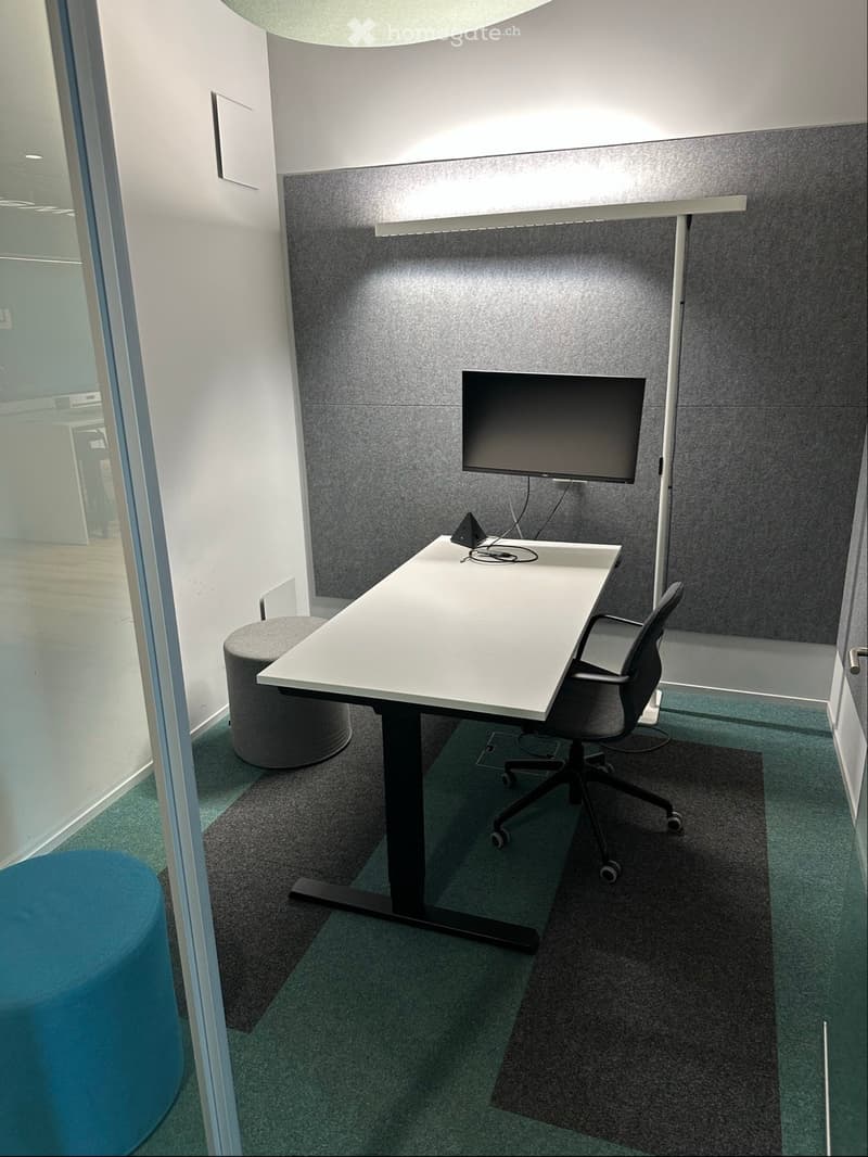 Small room, desk with monitor, gray office chair, gray carpet, white wall, gray ottoman, gray and blue stools, glass wall