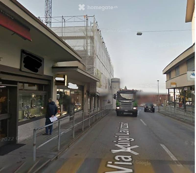 Street view showing a multi-story building with a covered walkway, a truck passing by, and a pedestrian on the sidewalk.