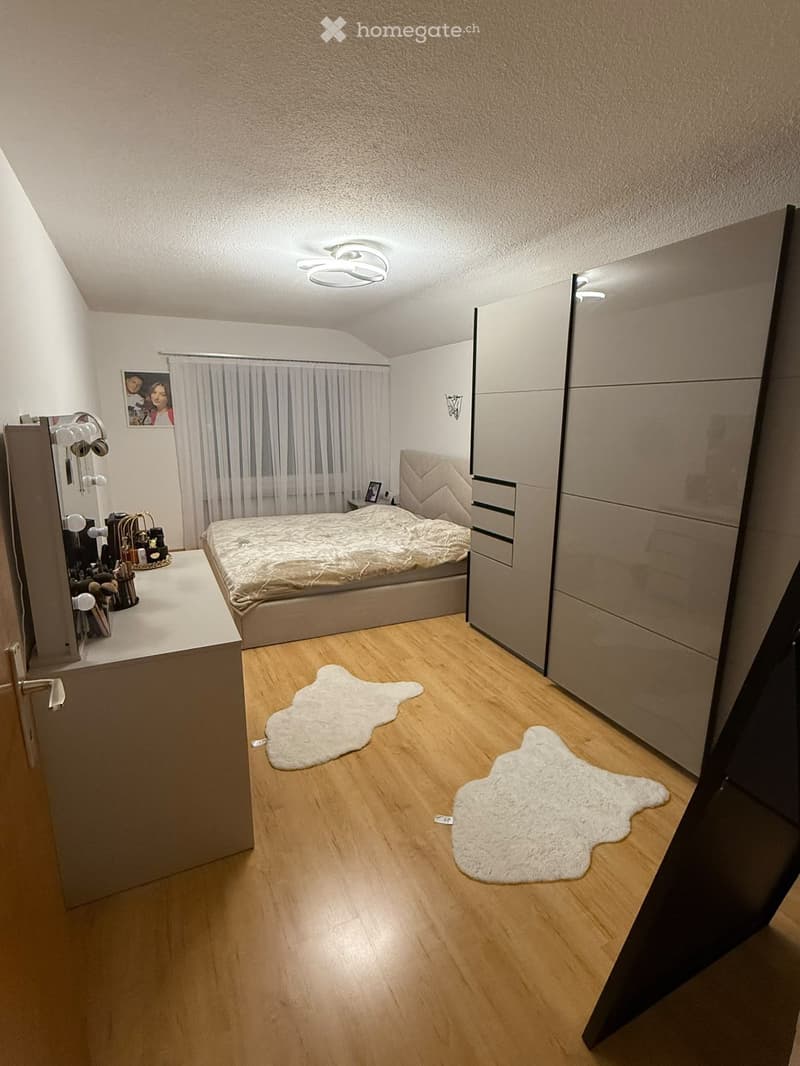 Modern bedroom with wooden flooring, a large bed with a textured headboard, a mirrored wardrobe, a vanity table with mirrors, and two white rugs on the floor.