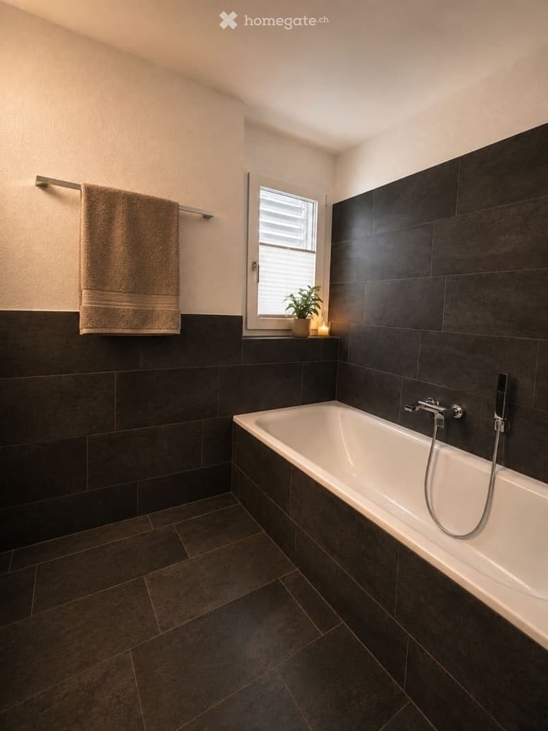 Modern bathroom with dark tiled walls and floor, a white bathtub, a towel on a rack, a small plant on the windowsill, and a candle.
