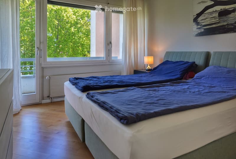 Bright bedroom with two single beds, wooden flooring, large windows with white curtains, and a nightstand with a lamp.