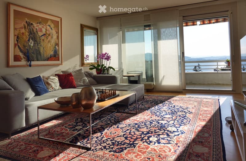 Modern living room with large windows offering a scenic view, featuring a grey sectional sofa, wooden coffee table with decorative items, chessboard, and a patterned rug.
