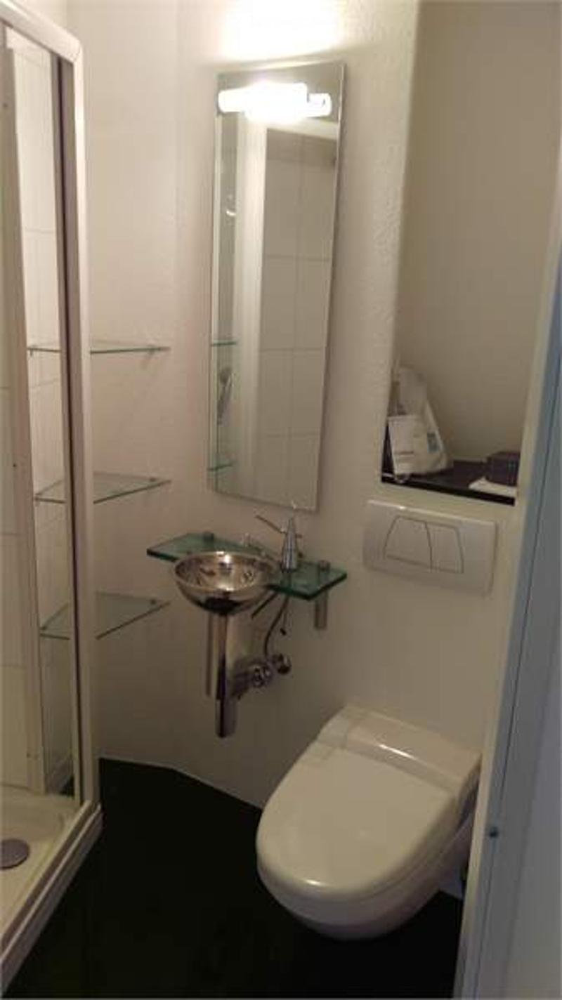 Compact bathroom with a toilet, sink, mirror, and shelves. The sink is mounted on a glass shelf, and the mirror is above it. The floor is dark, and the walls are white.