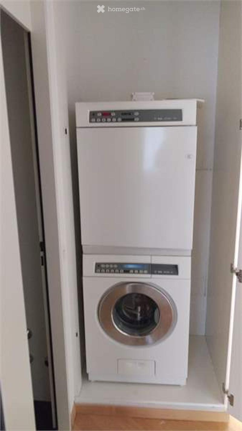 Compact bathroom with a stacked washing machine and dryer unit, white walls, wooden floor.