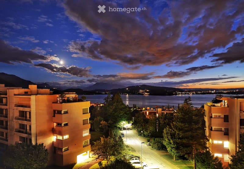 Nighttime view of a multi-story residential building with illuminated windows, surrounded by trees and parked cars, overlooking a lake and distant mountains.
