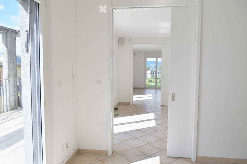 Bright, empty hallway with tiled flooring and white walls, leading to an open area with a view of a scenic landscape through large glass doors.
