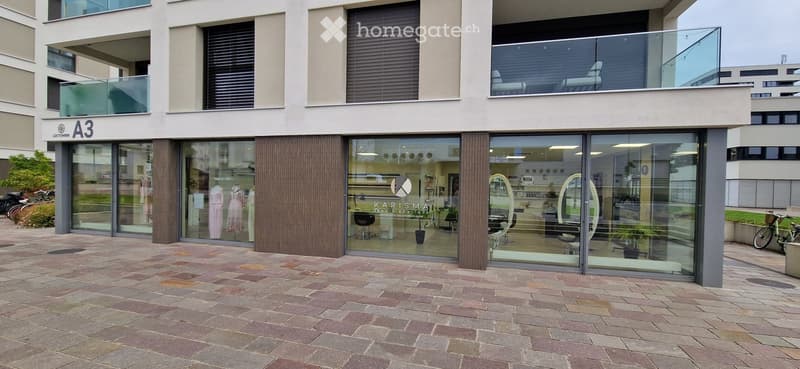 Modern building entrance with glass doors and a tiled pavement, featuring a beauty boutique with visible interior displays and a balcony above.