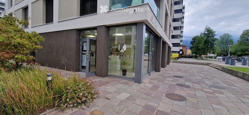 Modern building entrance with glass doors and a sign reading "KARISMA BEAUTY BOUTIQUE". The entrance features a paved walkway, small garden area with plants, and a modern design.