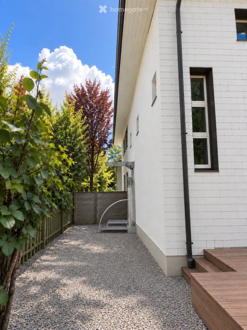 A modern garden area with a gravel pathway leading to a wooden deck, surrounded by greenery and a white house with large windows.