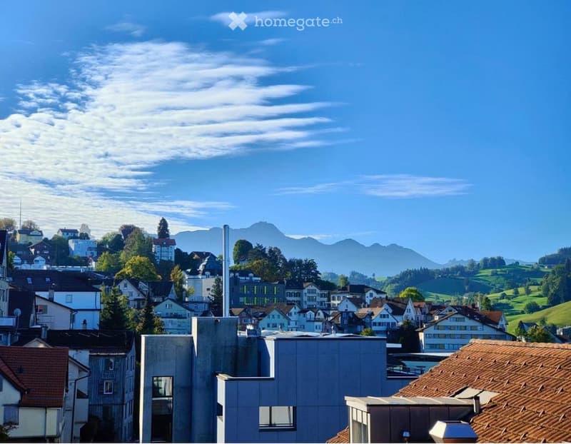 A panoramic view of a town with a mix of modern and traditional buildings, surrounded by green hills and distant mountains under a clear blue sky.