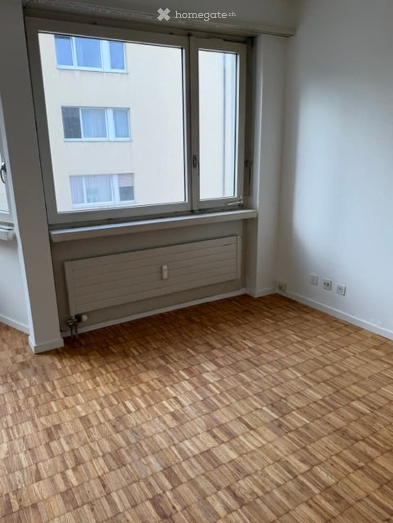 Empty room with wooden flooring, large window, radiator, and power outlets.