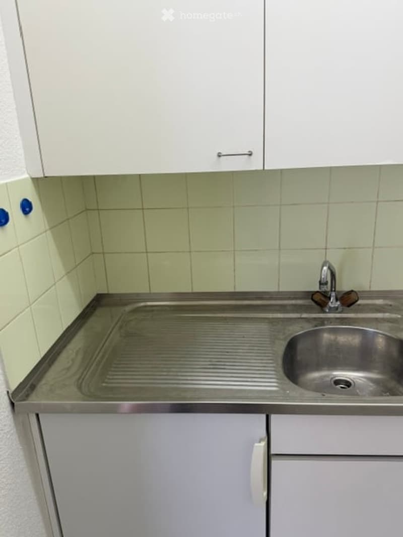 A compact kitchen with white cabinets, a stainless steel sink, and a faucet. The countertop is made of stainless steel, and the walls are tiled in a light green color.