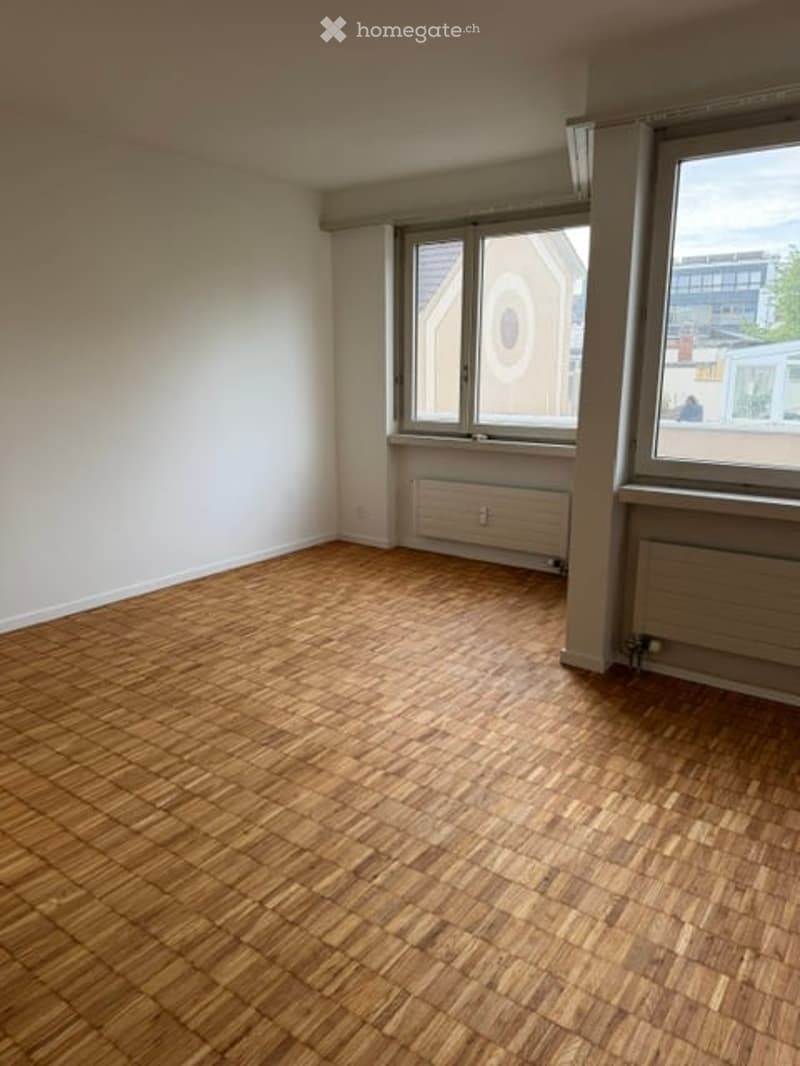 Empty room with wooden flooring, white walls, and large windows with radiators beneath them.