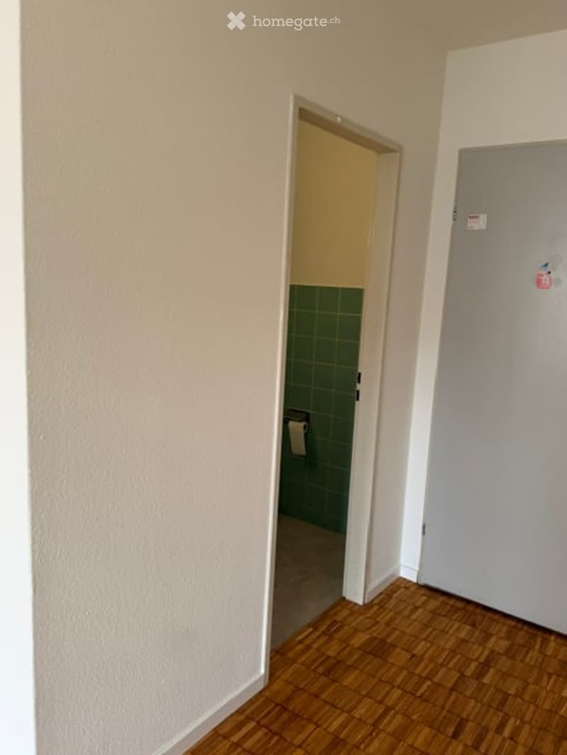Empty room with wooden flooring, white walls, and an open doorway leading to a small bathroom with green tiles.