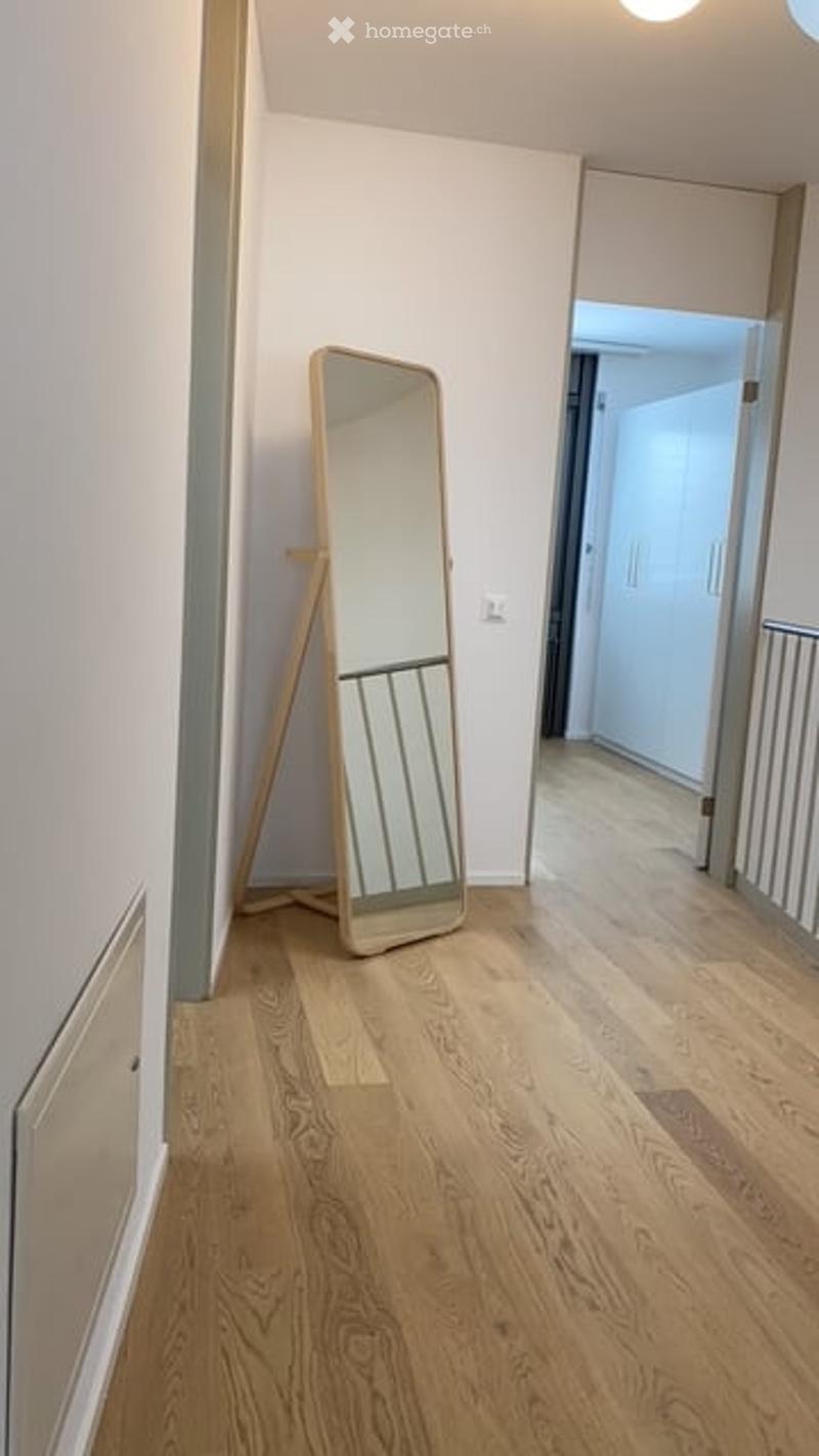 A clean, modern hallway with light wooden flooring, white walls, and a large floor mirror leaning against the wall. A doorway leads to another room, and a small railing is visible on the right side.