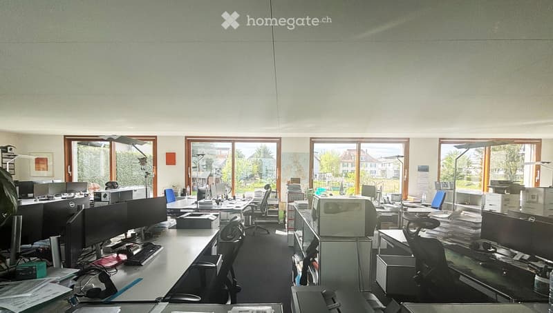 Office space with multiple desks, computer monitors, chairs, and office equipment. Large windows with views of the outside.