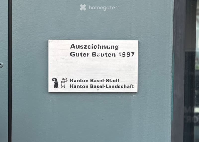 A silver sign with black text on a gray wall, reading 'Auszeichnung Guter Bauten 1997' and 'Kanton Basel-Stadt Kanton Basel-Landschaft'.